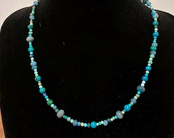 May include: A beaded necklace featuring a variety of round and faceted beads in shades of turquoise, teal, sky blue, and dark navy. The beads vary in size and texture, creating a multi-tonal blue and green color palette throughout the strand.