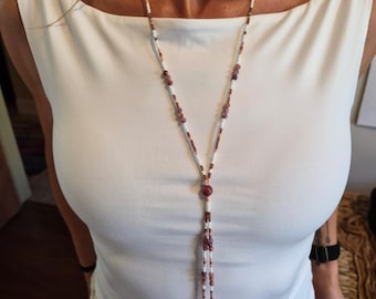 May include: A long lariat-style necklace featuring a delicate arrangement of small white, brown, and mauve beads. The design includes a central focal bead from which two strands descend, each finished with a single mauve bead at the bottom end.
