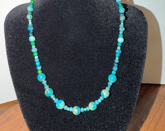 May include: A beaded necklace featuring a variety of round beads in shades of turquoise, teal, light blue, and green. The design incorporates larger, marbled spherical beads interspersed with smaller, translucent, and faceted beads.