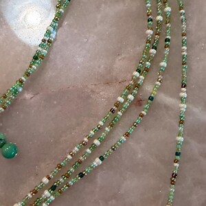 May include: A long beaded necklace featuring multiple strands of small glass beads in shades of light green, dark green, white, and metallic bronze. The necklace includes two larger, round, opaque turquoise-coloured beads at the end of the strands.