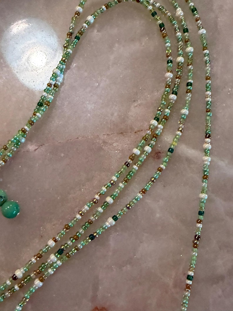May include: A long beaded necklace featuring multiple strands of small glass beads in shades of light green, dark green, white, and metallic bronze. The necklace includes two larger, round, opaque turquoise-coloured beads at the end of the strands.