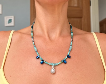 May include: A beaded necklace featuring a central silver-toned teardrop pendant encrusted with clear crystals. The strand consists of turquoise, light blue, and dark blue beads, interspersed with small metallic accents and four dark blue teardrop charms.