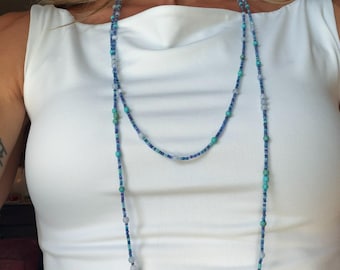 May include: A long beaded necklace featuring a variety of small round beads in shades of blue, turquoise, and translucent light gray. The necklace is strung in a continuous loop, allowing it to be worn as a single long strand or doubled over.