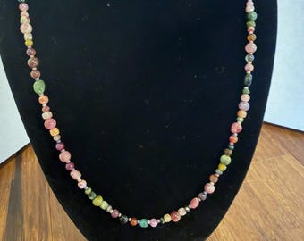 May include: A long necklace featuring a strand of small, irregularly shaped beads in various colors including shades of pink, green, brown, and translucent tones. The beads are strung together in a continuous loop, displaying a natural, varied texture and size throughout the entire length of the piece.