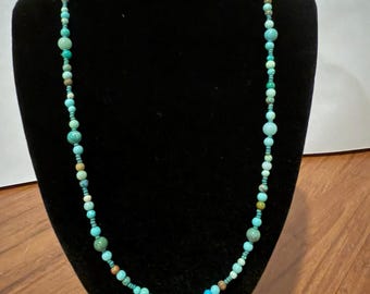 May include: A long necklace featuring a variety of round beads in shades of turquoise, light blue, and earthy brown. The beads vary in size and are strung together in a single strand, creating a multi-textured appearance with natural color variations.