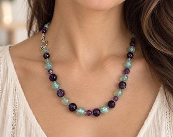 May include: A beaded necklace featuring alternating round, polished deep purple beads and translucent light green beads. The necklace is secured with a silver-toned metal hook-and-eye clasp, creating a continuous circular pattern around the neck.