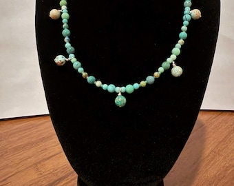 May include: A beaded necklace featuring a strand of small, round, turquoise and green-toned beads. The necklace is accented with four larger, dangling, spherical beads in mottled shades of tan, brown, and teal, each secured with silver-toned wire.