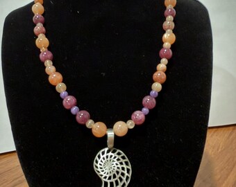 May include: A beaded necklace featuring alternating round peach, light orange, and deep purple gemstone beads. The necklace includes a silver-toned metal pendant shaped like a nautilus shell with a detailed spiral cutout design.