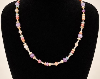 May include: A beaded necklace featuring a variety of round and disc-shaped beads in shades of lavender, peach, cream, and soft green. The beads are interspersed with small metallic spacers, creating a multi-colored, textured design.
