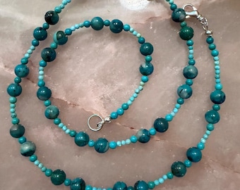 May include: A beaded necklace featuring alternating sizes of round turquoise-colored stones in shades of teal and light blue. The strand is accented with small, uniform light blue beads and secured with a silver-toned metal lobster claw clasp.