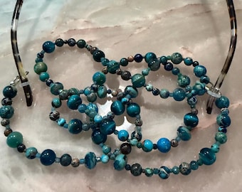 May include: A beaded eyeglass chain featuring a variety of round, polished stones in shades of teal, turquoise, and deep blue. The chain is attached to the temples of a pair of tortoiseshell-patterned eyeglasses with clear rubber loops.