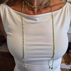 May include: A long, multi-strand beaded necklace featuring small glass beads in various shades of green, white, and metallic gold. The necklace wraps around the neck and hangs down in two long strands, each finished with a larger green bead and silver hardware.