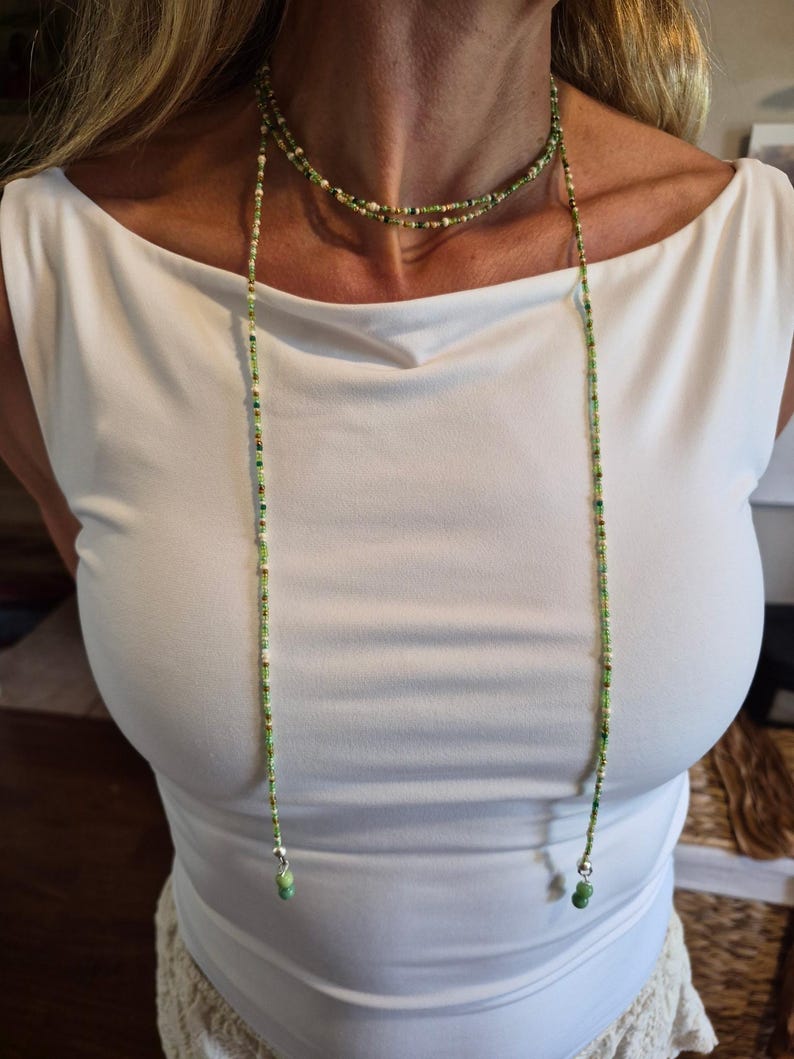 May include: A long, multi-strand beaded necklace featuring small glass beads in various shades of green, white, and metallic gold. The necklace wraps around the neck and hangs down in two long strands, each finished with a larger green bead and silver hardware.