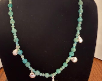 May include: A beaded necklace featuring round, light green-blue stones of varying sizes. The necklace is accented with four silver-toned charms, including three small seashell shapes and one central starfish charm hanging from the strand.