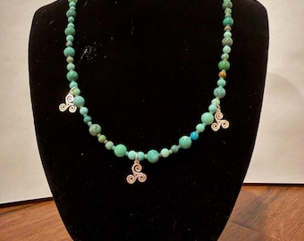 May include: A beaded necklace featuring round turquoise-colored stones in varying shades of green and blue. The necklace is accented with three silver-toned metal triskele charms spaced evenly along the strand of beads.