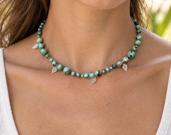 May include: A beaded necklace featuring round turquoise-colored stones with dark matrix patterns, interspersed with small silver-toned metal leaf charms. The beads vary in size, creating a graduated look around the neck, secured with a clasp.