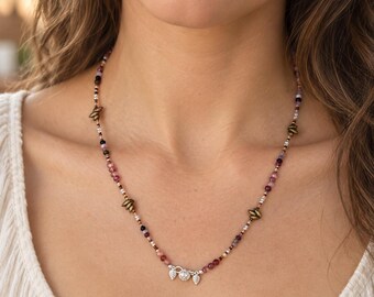 May include: A beaded necklace featuring small, multi-colored translucent beads in shades of pink, purple, and clear, interspersed with metallic bronze-toned leaf-shaped accents. The center features a silver-toned charm with three small pendants.