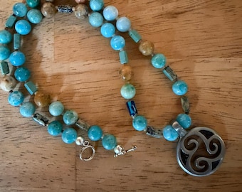 May include: A beaded necklace featuring a variety of round and cylindrical beads in shades of turquoise, blue, brown, and tan. The necklace includes a silver-toned metal pendant with a circular triskelion cutout design and a toggle clasp closure.