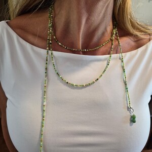 May include: A long lariat necklace featuring a delicate strand of small green, white, and brown seed beads. The necklace wraps around the neck and hangs down with two silver-toned metal findings, each finished with a pair of smooth green beads.