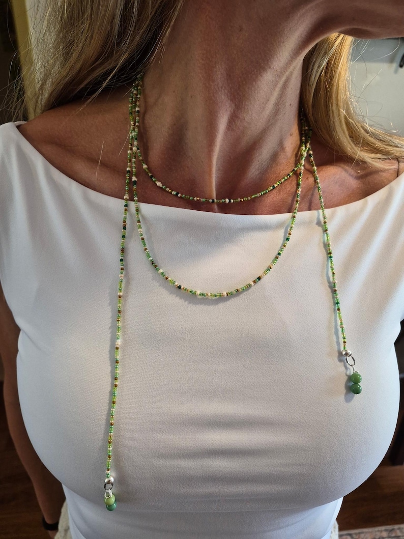 May include: A long lariat necklace featuring a delicate strand of small green, white, and brown seed beads. The necklace wraps around the neck and hangs down with two silver-toned metal findings, each finished with a pair of smooth green beads.