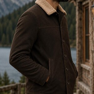 Puede incluir: Chaqueta marr&oacute;n oscuro con cuello de borreguillo beige. La chaqueta tiene cierre de botones, dos bolsillos delanteros y un dise&ntilde;o cl&aacute;sico. La persona est&aacute; de pie afuera cerca de un lago y un edificio de madera.
