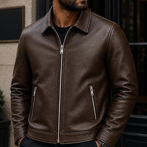 Puede incluir: Una chaqueta de cuero marrón oscuro con cuello y cremallera completa. La chaqueta tiene dos bolsillos con cremallera y una superficie texturizada. Las mangas son largas y la chaqueta parece estar bien ajustada. El estilo general es clásico y versátil.