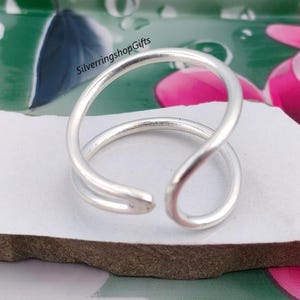 May include: A silver-coloured open ring featuring a smooth, rounded band that curves to create a double-loop design. The metal has a polished finish, and the text SilverringshopGifts is visible in black lettering near the top of the ring structure.