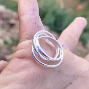 Puede incluir: Un anillo de color plateado compuesto por tres bandas circulares, lisas y entrelazadas. El metal tiene un acabado pulido y reflectante, y las tres bandas separadas están unidas para formar una sola pieza de joyería coherente.