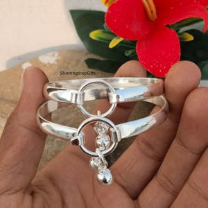 May include: Two silver bangles with a connecting ring and dangling charms. The bangles are held in a hand, with a red flower and green leaves in the background. The text "SilverringshopGifts" is visible.