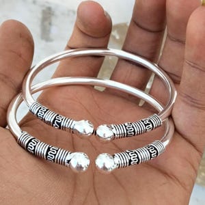 Puede incluir: Dos brazaletes abiertos de tono plateado con acabado pulido. Cada brazalete tiene terminales redondeados y lisos, y una sección decorativa cerca de los extremos caracterizada por intrincados patrones grabados oscuros y detalles acanalados.