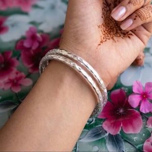 May include: Two silver-toned bangles with a hammered, textured surface. The bracelets feature a rounded, polished finish that reflects light, creating a series of small, irregular indentations across the entire circumference of each metal band.