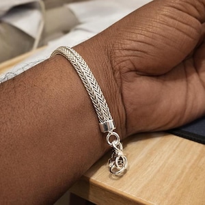 May include: A silver bracelet with a woven chain design is clasped on a wrist. The bracelet has a textured, braided appearance and a secure clasp. The jewelry is displayed on a wooden surface.