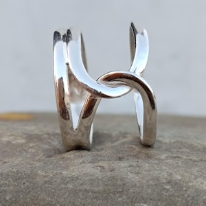 Puede incluir: Primer plano de un anillo plateado con un diseño único. El anillo presenta un diseño abierto y entrelazado con un acabado pulido. Se muestra sobre una superficie gris texturizada, resaltando sus cualidades reflectantes. El diseño del anillo es moderno y minimalista.