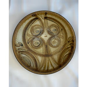 May include: A round, ceramic plate with a brown and beige color scheme. The plate features a central design of swirling patterns and lines, with a dark brown rim. The design is symmetrical and appears to be hand-painted.
