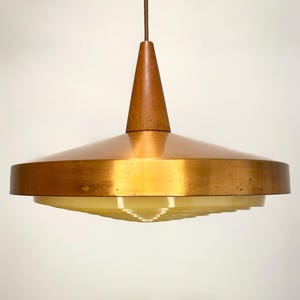 Peut inclure: Un luminaire suspendu de style rétro. Il présente un abat-jour en forme de soucoupe de couleur cuivrée, surmonté d'un cône en bois. La partie inférieure de l'abat-jour est jaune clair. Il est suspendu par un câble marron.