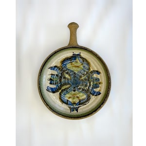 Puede incluir: Plato de cerámica redondo con asa. El plato presenta un diseño abstracto azul y verde central sobre un fondo crema, enmarcado por un borde verde y marrón. El asa es de color marrón claro.