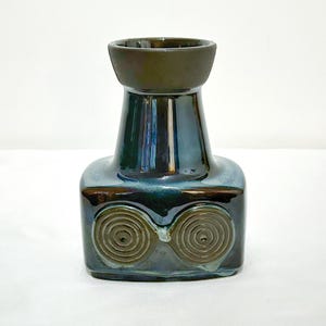 Peut inclure: Un vase en céramique avec un bord marron foncé et un corps bleu sarcelle. Le vase a une base carrée avec deux motifs en spirale et un col cylindrique. Le vernis a une finition brillante.