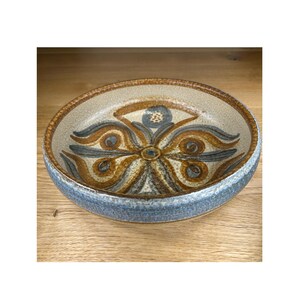 May include: A ceramic bowl with a floral design. The bowl has a light beige interior with brown, blue, and gray floral patterns. The rim is a darker blue, and the exterior is a light beige color. The bowl is round and appears to be handmade.