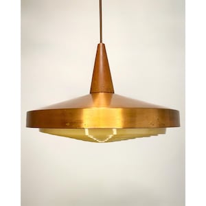 Könnte beinhalten: Eine Hängeleuchte im Retro-Stil. Sie hat einen konischen Holzaufsatz, einen breiten, kreisförmigen kupferfarbenen Schirm und einen geschichteten, gelblich getönten Glasdiffusor. Die Leuchte hängt an einem braunen Kabel.