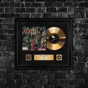 Op de afbeelding: Gegoten gouden plaat en albumhoes van "Reign in Blood" van Slayer. De albumhoes heeft een donkere, gedetailleerde illustratie. De gouden plaat wordt naast de albumhoes weergegeven, met een zwarte lijst en een gouden plaat.