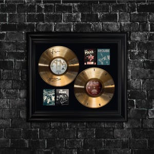 May include: Framed gold record display featuring two gold records and four album covers. The records are signed and labeled. The album covers are for the band The Police. The frame is black and set against a black brick wall.