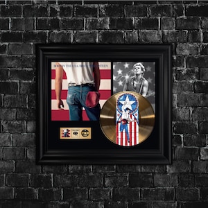 May include: Framed display featuring a gold record and two images. The gold record has a star and stripes design. One image shows a person in front of an American flag. The other image is a black and white photo of a person playing a guitar.