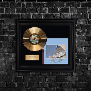 Könnte beinhalten: Gerahmte goldene Schallplatte und Albumcover von Dire Straits' "Brothers in Arms". Die goldene Schallplatte wird zusammen mit dem Albumcover mit einer Gitarrenillustration und Unterschriften präsentiert. Der Rahmen ist schwarz, vor einer dunklen Backsteinmauer.