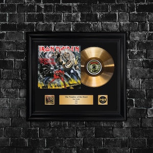 Puede incluir: Disco de oro enmarcado del álbum "The Number of the Beast" de Iron Maiden. El marco incluye la portada del álbum, un disco de oro y una placa con el título del álbum y el nombre de la banda. El marco es negro.