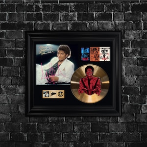 Op de afbeelding: Een ingelijste presentatie met Michael Jacksons "Thriller" albumkunst en een gouden plaat. De lijst bevat een foto van Michael Jackson, albumhoezen en een gouden plaat met een zombie-afbeelding. De lijst is zwart en gemonteerd op een donkere bakstenen muur.