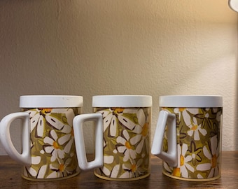 May include: Three ceramic mugs with white handles and lids. The mugs feature a floral pattern of white daisies on a green and brown background. The mugs are cylindrical and sit on a wooden surface.