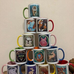 May include: A pyramid of white ceramic mugs, each adorned with a different animal illustration from the Chinese zodiac. The mugs have colored handles and rims, including green, red, yellow, and blue. The mugs are stacked on top of each other.
