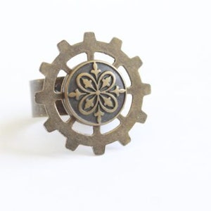 May include: A bronze-colored ring with a gear-like design. The ring features a central circular element with a floral pattern. The ring is set against a white background, highlighting its intricate details and steampunk aesthetic.