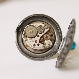 May include: A vintage, open locket ring with a silver-tone finish. The locket reveals the intricate gears and jewels of a watch mechanism. A small, turquoise-colored stone adorns the side of the locket.