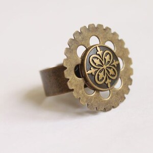 May include: A steampunk-style ring with a bronze-colored gear design. The ring features a central, circular element with a floral pattern. The band and gear are a matching bronze color, creating a unique, industrial aesthetic.